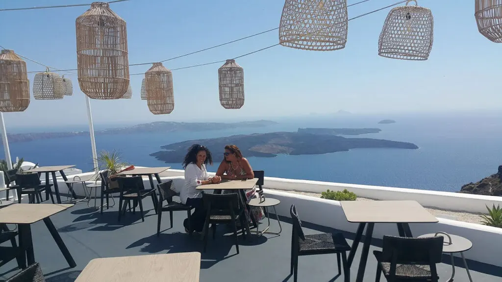 two women sit on the restaurant terrace in santorini overlooking the sea and the famous santorini volcano