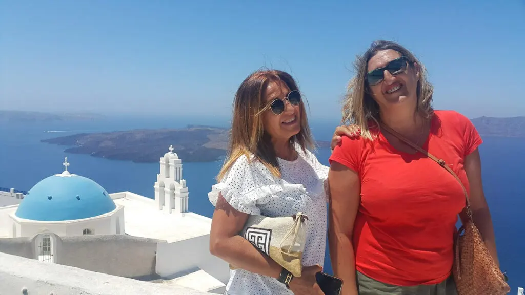 two women pose at the top of a church in santorini overlooking the sea